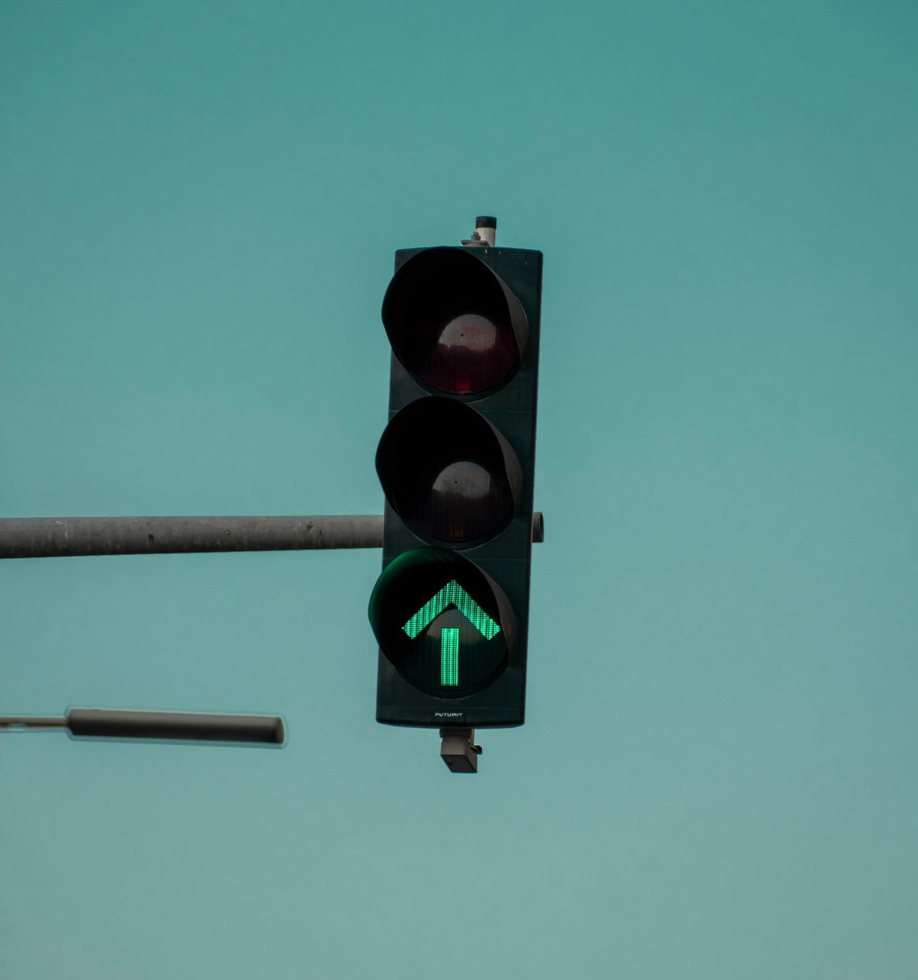 Traffic light with a green arrow illuminated against a light teal sky, indicating permission to proceed straight.